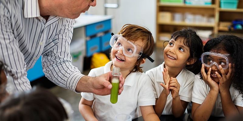 Elementary school children engaged in an experiment with a teacher holding a beaker with green liquid.