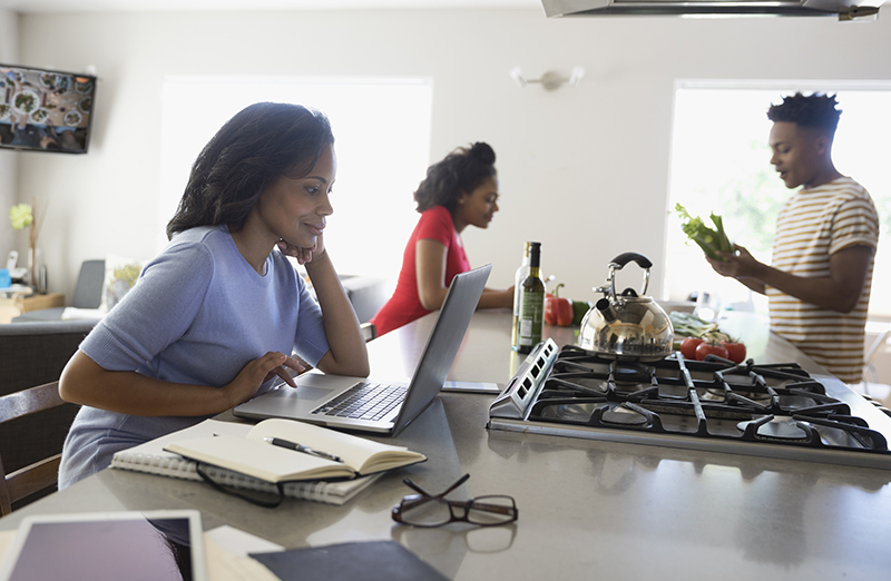 African American mother working at laptop and teenage children cooking in kitchen