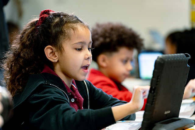 Student uses a tablet in a classroom, with another student working in the background