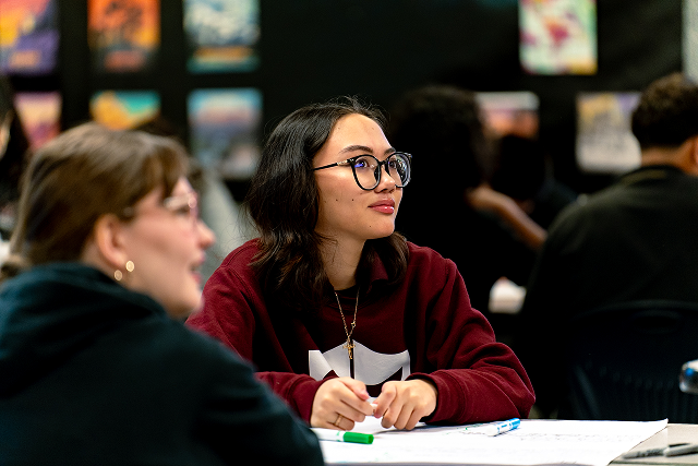 Student wearing glasses sits at a table in a classroom, listening and taking notes during a discussion