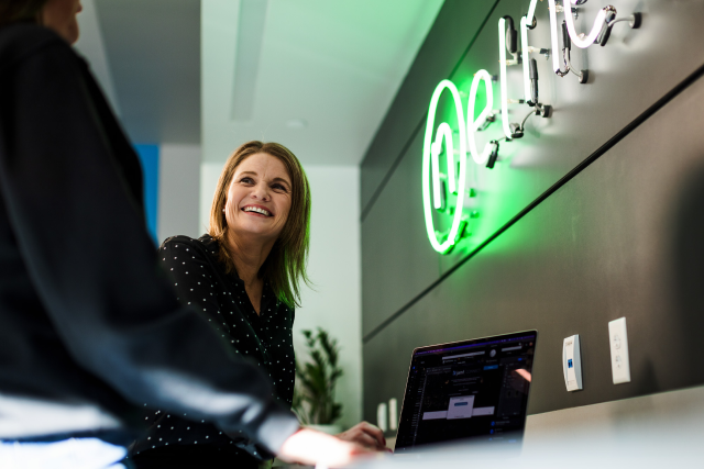 Smiling employee talks with a colleague at a desk with a laptop, with a green neon sign on the wall behind them