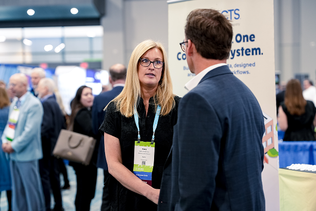 Two conference attendees talk at a trade show booth, with a FACTS banner in the background and other attendees nearby.