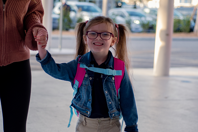 Smiling child wearing glasses and a backpack holds an adult’s hand while walking outside near a school entrance.