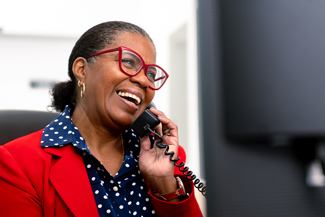 Smiling woman wearing red glasses and a red blazer talks on a desk phone in an office