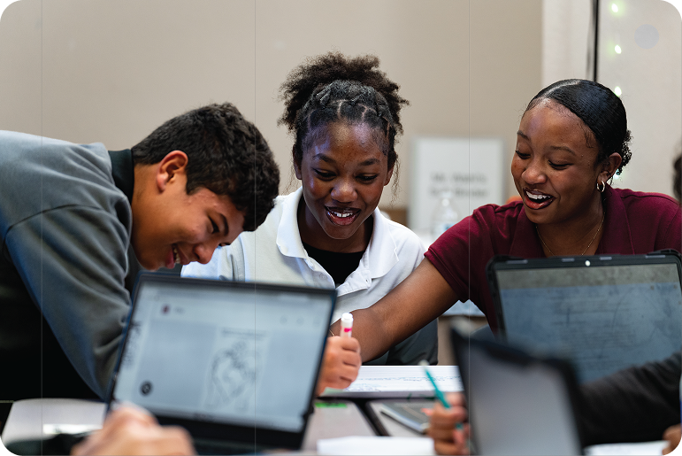 Students work together at a table with laptops and notebooks, smiling during a group activity