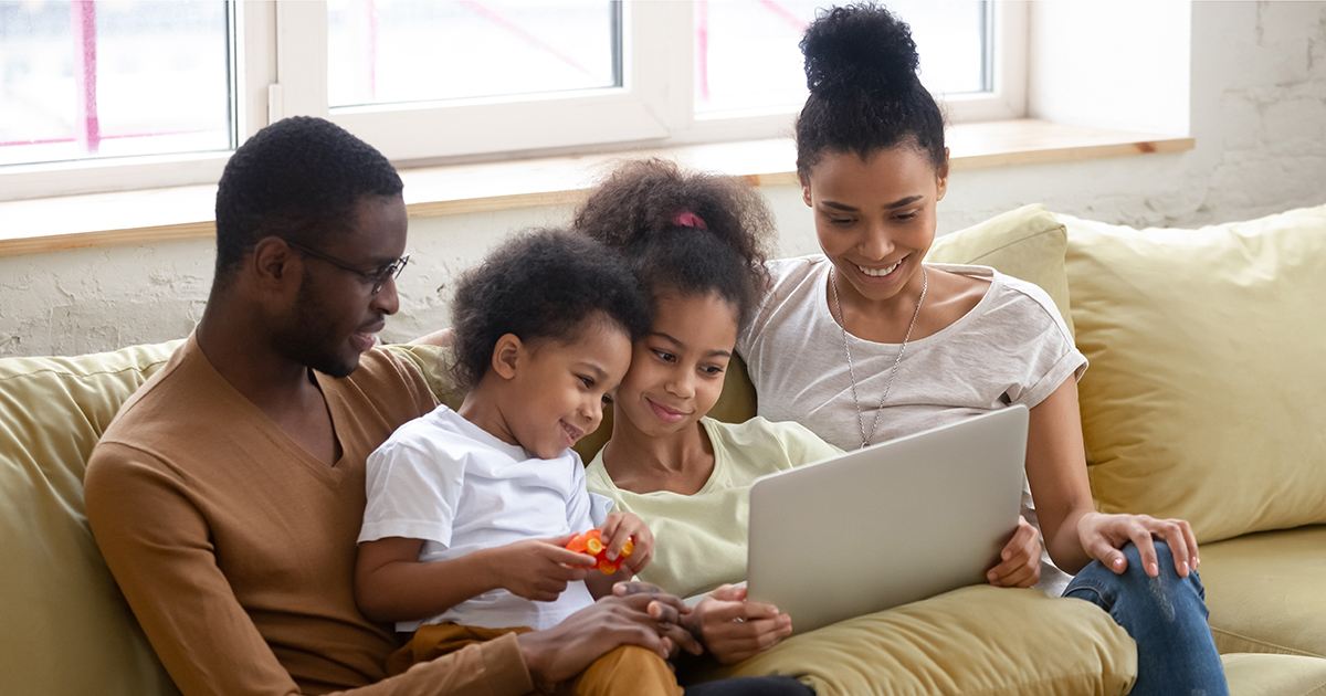 A father, mother, and two young children sit together on a couch looking at a laptop.