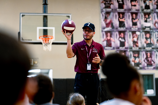 Coach or teacher holds a football while speaking to students in a gymnasium, with a basketball hoop in the background