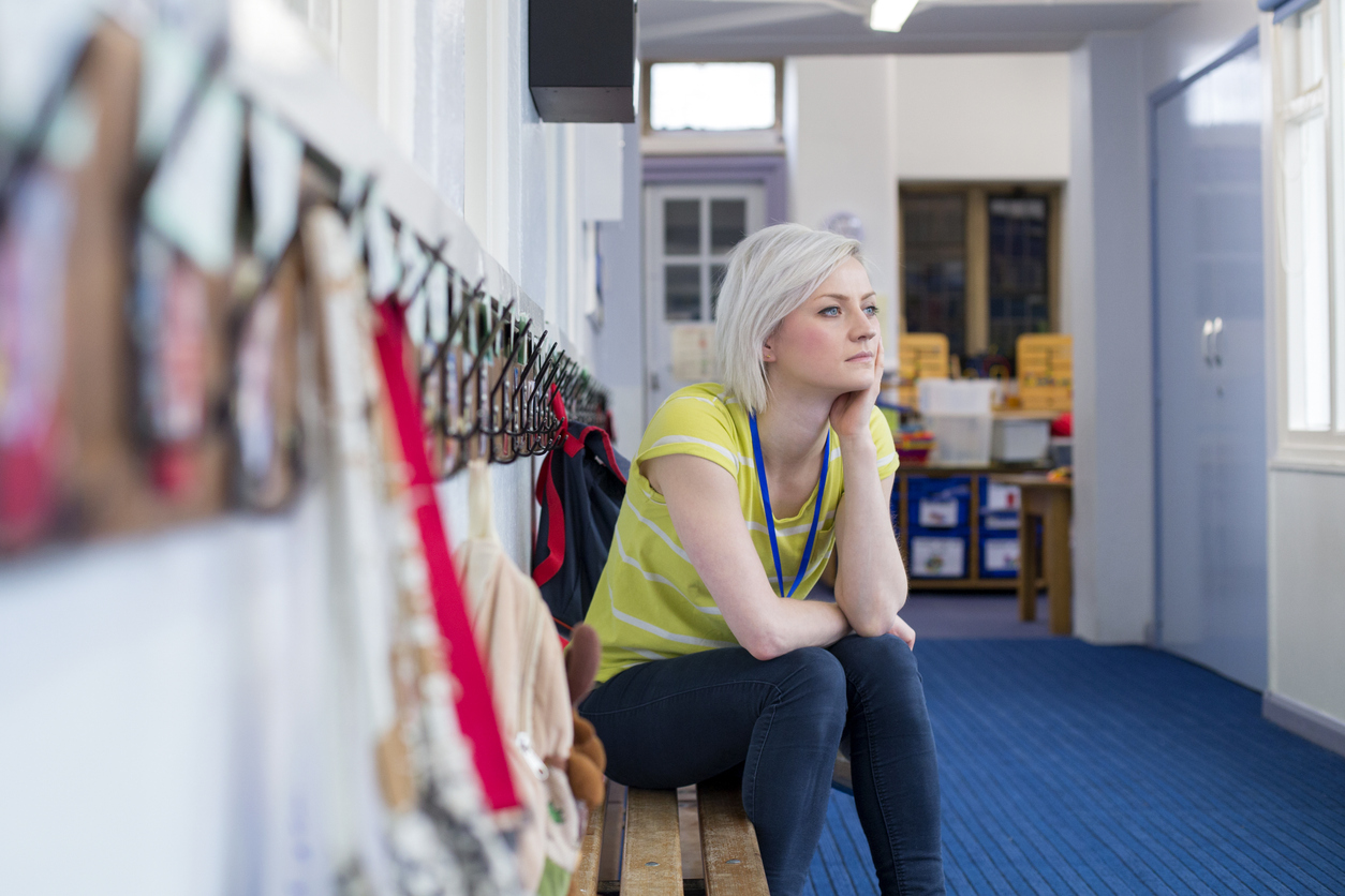 A young teacher sitting on a bench in the cloakroom of school. She is looking pensively out of the window.