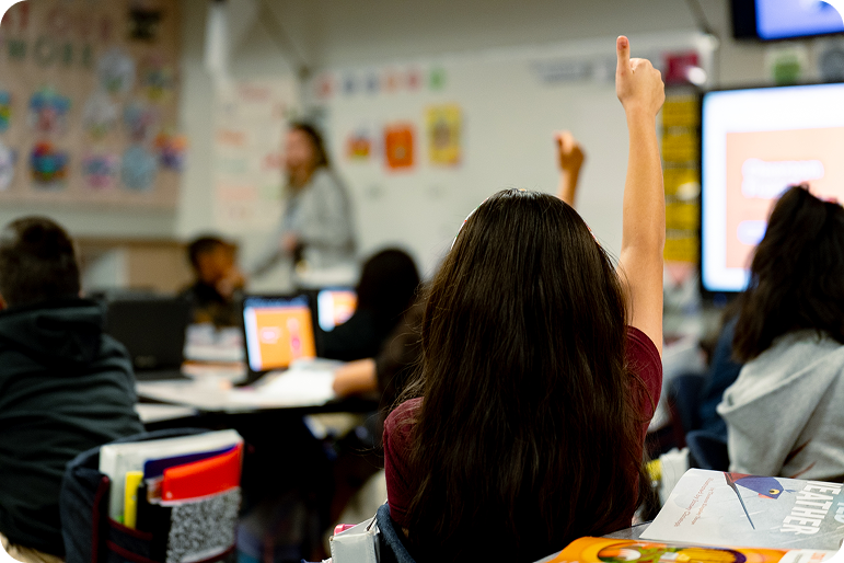 Student raises a hand in a classroom while a teacher presents at the front
