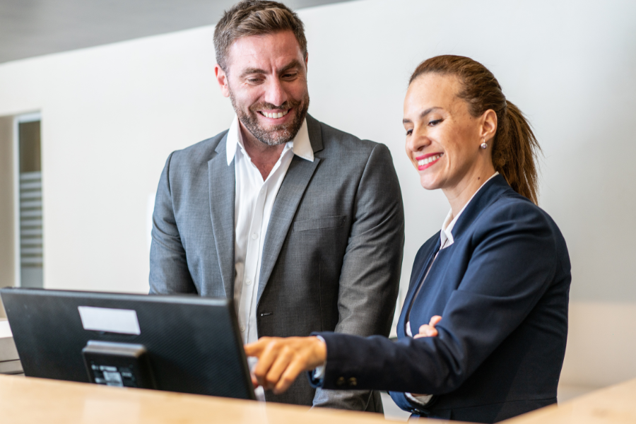 Man and woman in a professional setting, standing and smiling, looking at something together on a desktop monitor